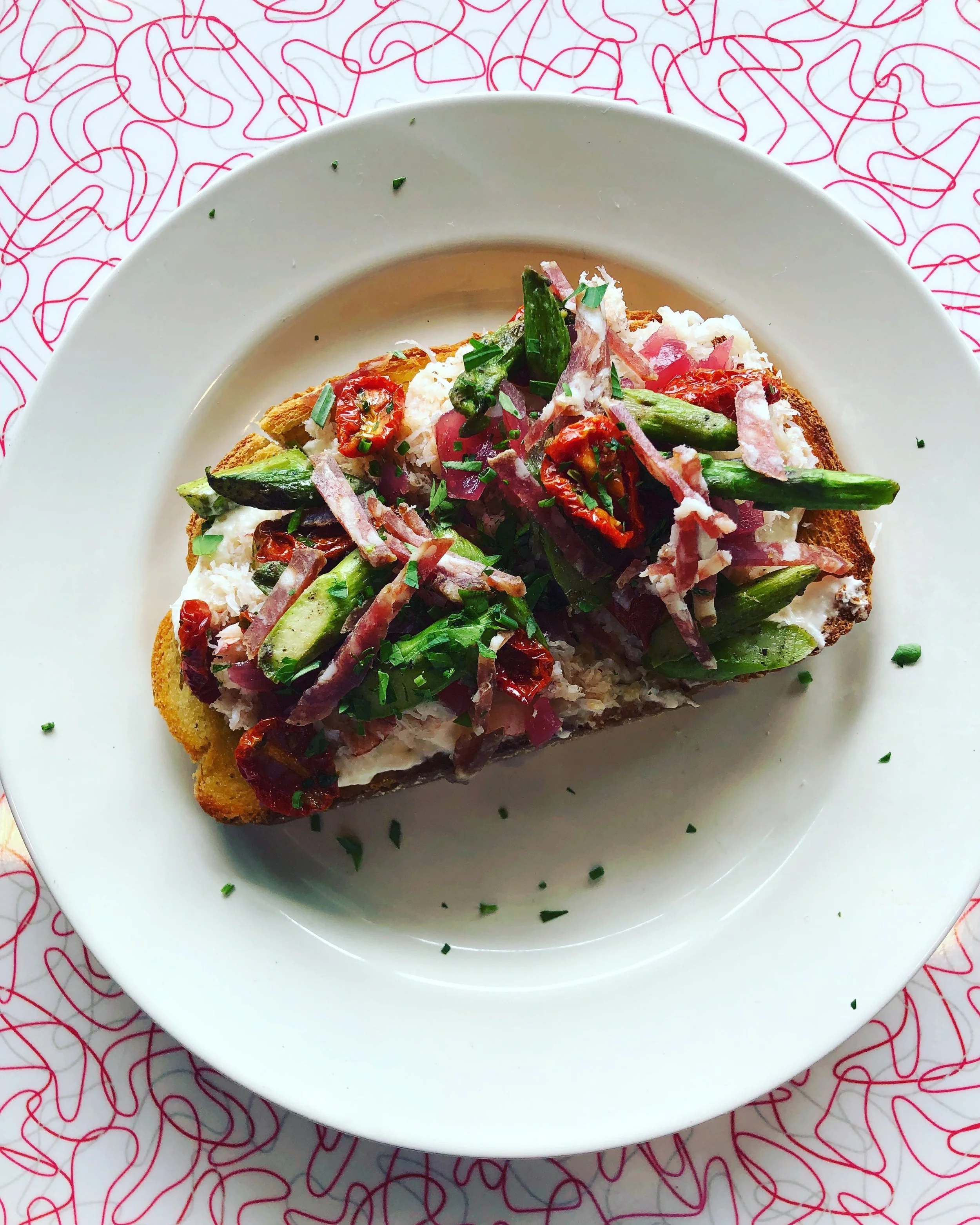 Toast topped with a white spread, asparagus, tomatoes, red onion, and herbs on a white plate