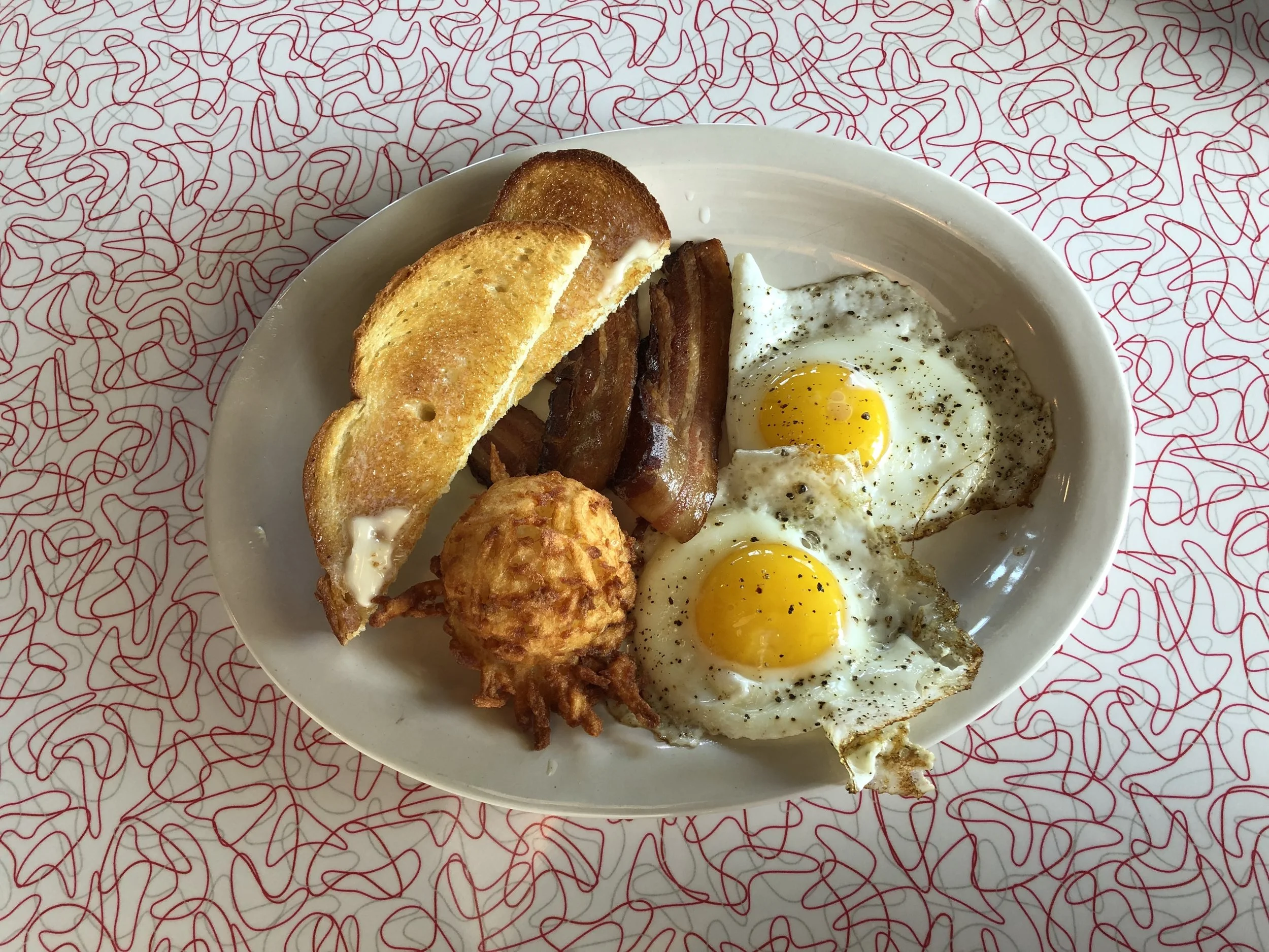 Two sunny-side-up eggs with bacon, buttered toast, and a golden potato side on a white plate
