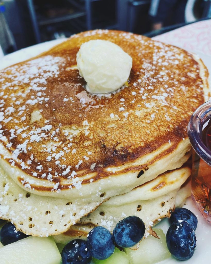 Close-up of pancakes topped with butter and powdered sugar beside blueberries and syrup